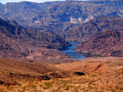 [Zoomed out view of the prior photo showing the massive amounts of very dark brown rock surrounding the sliver of water. No vegetation is visible on the rock, but some is visible on the sandy colored dirt in the foreground.]